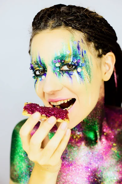 Nahaufnahme Porträt eines schönen Mädchens mit kreativem Glitzer-Make-up beim Essen eines Snacks, der mit Glitzer bedeckt ist — Stockfoto