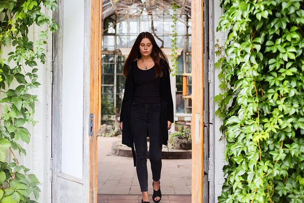 Potret gaya hidup gadis mengenakan kaos hitam kosong, celana jeans dan mantel berpose terhadap bangunan ditutupi dengan daun hijau — Stok Foto