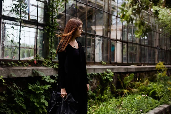 Портрет девушки, одетой в пустую черную футболку, джинсы и пальто, позирующую против здания, покрытого зелеными листьями — стоковое фото