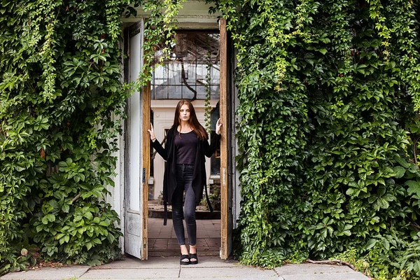 라이프 스타일 초상화 빈 블랙 t-셔츠, 청바지와 건물에 대 한 포즈 코트를 입고 여자의 녹색 잎으로 덮여 — 스톡 사진