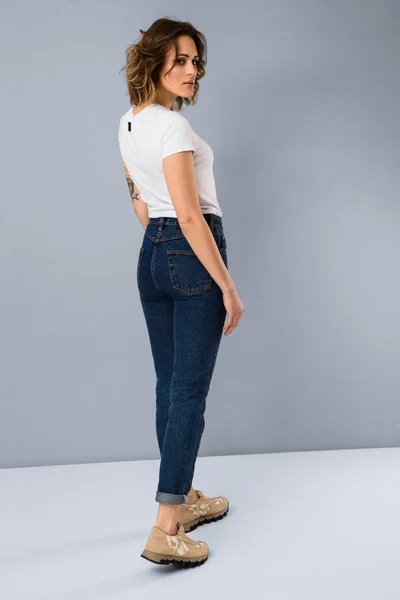 Portrait of stylish young girl in basic white t-shirt and high waisted blue jeans  posing in studio on grey background