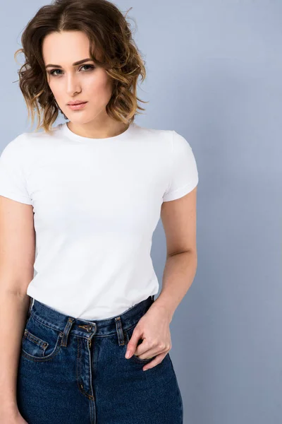 Portrait of stylish young girl in basic white t-shirt and high waisted blue jeans  posing in studio on grey background