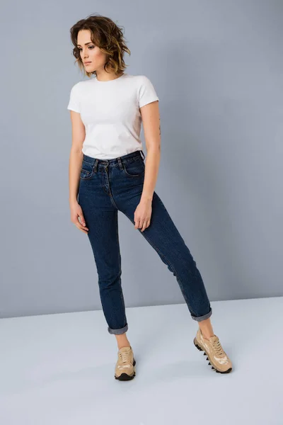 Portrait of stylish young girl in basic white t-shirt and high waisted blue jeans  posing in studio on grey background