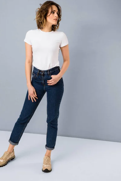 Portrait of stylish young girl in basic white t-shirt and high waisted blue jeans  posing in studio on grey background