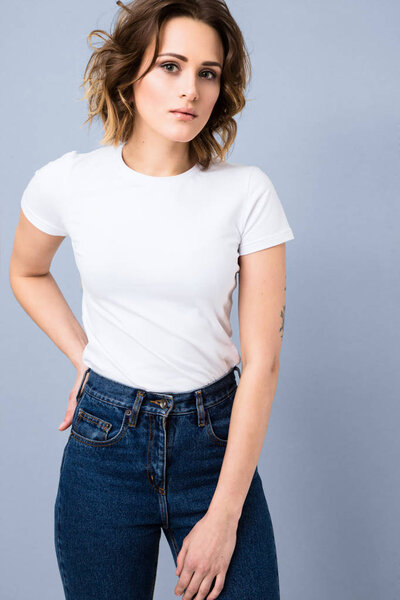 Portrait of stylish young girl in basic white t-shirt and high waisted blue jeans  posing in studio on grey background