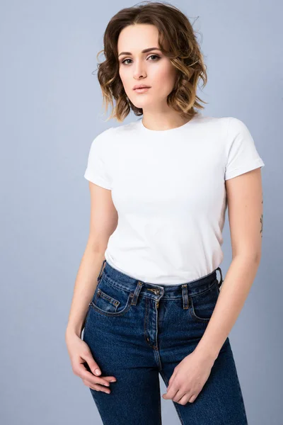 Portrait of stylish young girl in basic white t-shirt and high waisted blue jeans  posing in studio on grey background