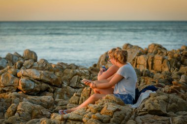 Gün batımında kayaların üzerinde oturan iki genç kadın, Agnes Water, Queensland, Avustralya