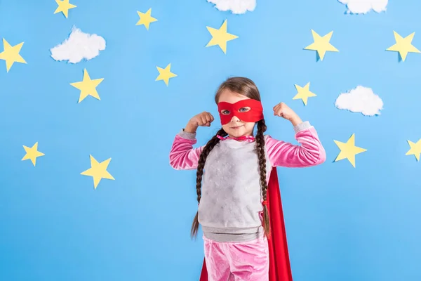 Ragazza bionda di sei anni vestita da supereroe che si diverte a casa. Ragazzo sullo sfondo della parete blu brillante . — Foto Stock