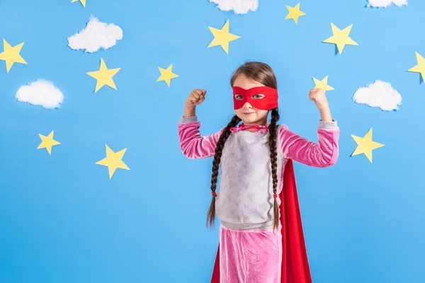 Ragazza bionda di sei anni vestita da supereroe che si diverte a casa. Ragazzo sullo sfondo della parete blu brillante . — Foto Stock