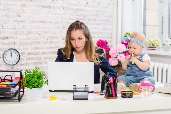 Joven y alegre mujer de negocios hermosa mirando a la computadora portátil mientras está sentada en su lugar de trabajo con su hija pequeña — Foto de Stock