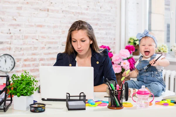 Веселая молодая красивая деловая женщина смотрит на ноутбук, сидя на своем рабочем месте со своей маленькой дочерью — стоковое фото