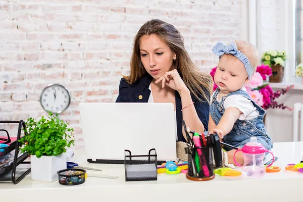 Jovem e alegre mulher de negócios bonita olhando para o laptop enquanto se senta em seu local de trabalho com sua filhinha — Fotografia de Stock