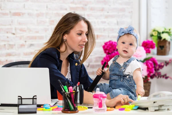 Jovem e alegre mulher de negócios bonita olhando para o telefone enquanto se senta em seu local de trabalho com sua filhinha — Fotografia de Stock