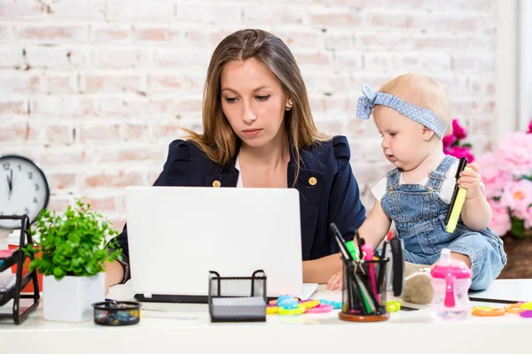 Весела молода красива бізнес-леді дивиться на ноутбук, сидячи на своєму робочому місці зі своєю маленькою донькою — стокове фото