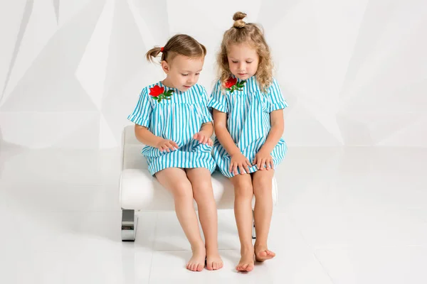 Dvě malé kamarádky ve stejném tyrkysové šaty sedí na židli v ateliéru s bílými stěnami — Stock fotografie