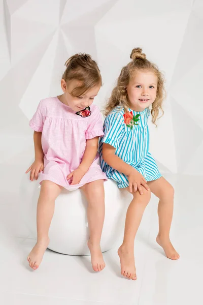 Twee kleine meisjes in de identieke jurken van verschillende kleuren, zittend op een stoel in een studio met witte muren — Stockfoto
