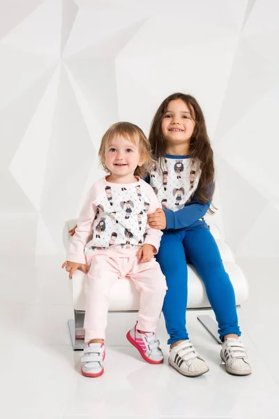 Две стильные маленькие девочки сидят на круглом стуле на белом фоне стены — стоковое фото