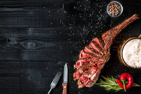 Grill száraz éves borda, marhahús, fűszer, zöldségek és egy pohár világos sör közeli kép a fekete fából készült háttér — Stock Fotó
