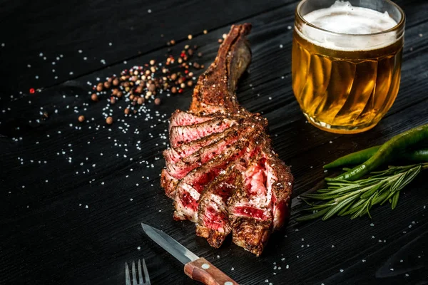 Grill trocken gereifte Rinderrippe mit Gewürzen, Gemüse und einem Glas leichtem Bier in Großaufnahme auf schwarzem Holzhintergrund — Stockfoto