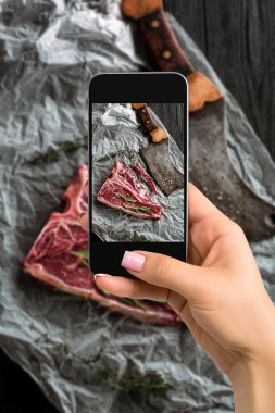 Photographing food concept - woman takes picture of raw dry aged t-bone steaks for grill with fresh herbs and cleaver clipart