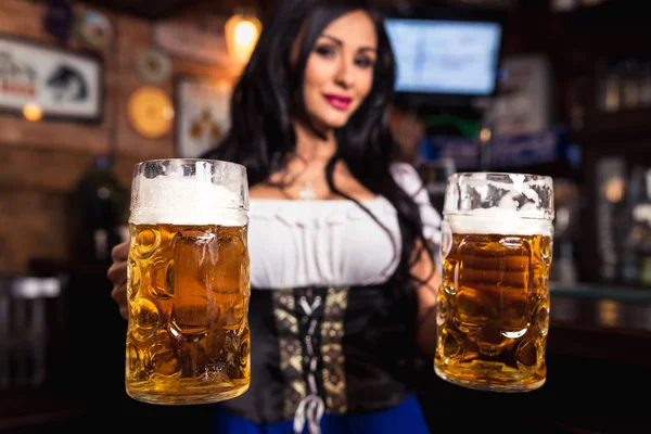 Joven camarera sexy Oktoberfest, con un vestido tradicional bávaro, sirviendo tazas de cerveza grandes en el bar . — Foto de Stock