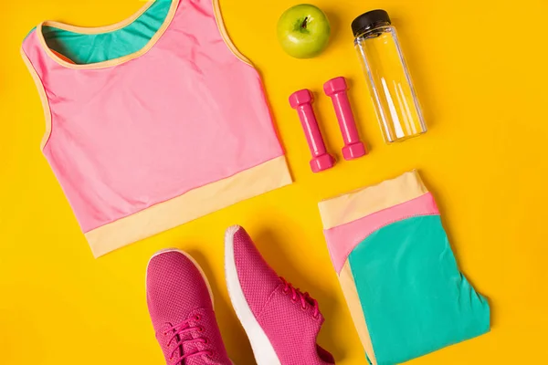 Fitness tartozékok sárga háttéren. Cipők, üveg víz, súlyzók és sport top. — Stock Fotó