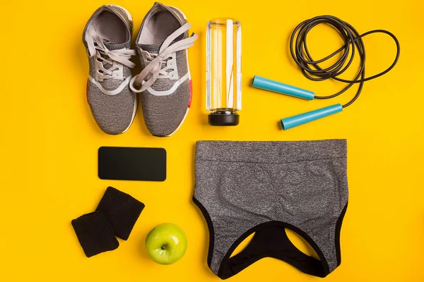 Fitness accessories on a yellow background. Sneakers, bottle of water, apple and sport top.