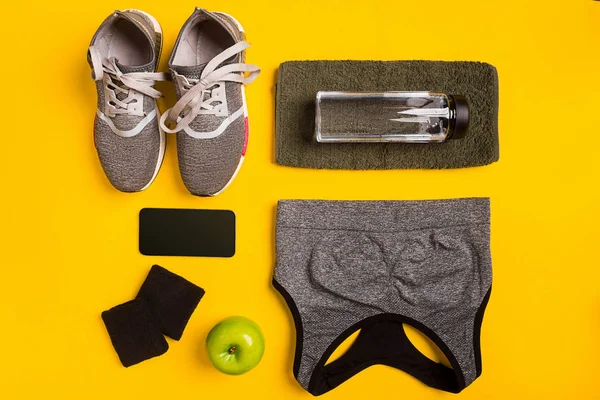 Fitness tartozékok sárga háttéren. Cipők, üveg víz, intelligens, törölköző és sport top. — Stock Fotó