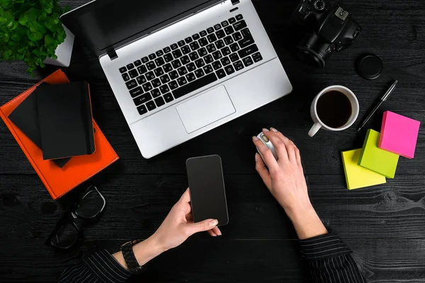 Frauenhände halten einen Smart und tippen auf der Tastatur eines Laptops auf einem schwarzen Holztisch. — Stockfoto