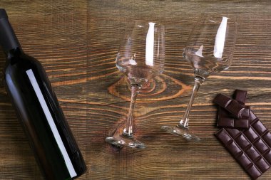 Bar of chocolate, bottle and two glasses of wine on wooden background from top view clipart