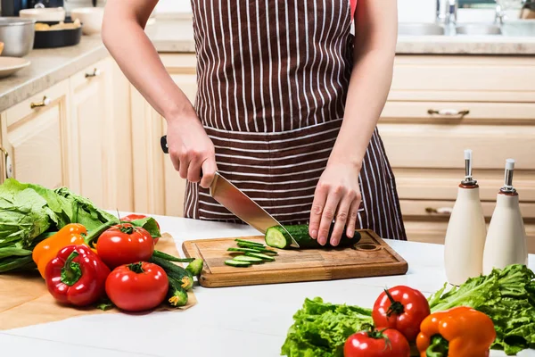 Mujer joven cocinando en la cocina en casa. Una mujer corta un pepino y verduras con un cuchillo . — Foto de Stock