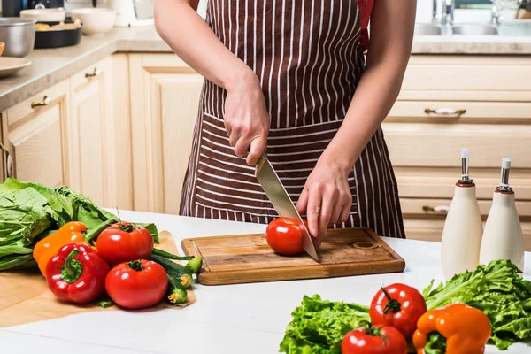 Mujer joven cocinando en la cocina en casa. Una mujer corta un tomate y verduras con un cuchillo . — Foto de Stock