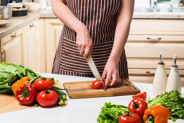 Mujer joven cocinando en la cocina en casa. Una mujer corta un tomate y verduras con un cuchillo . — Foto de Stock