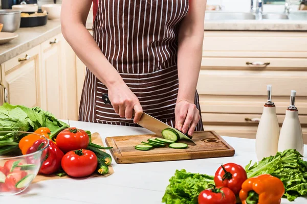 Mujer joven cocinando en la cocina en casa. Una mujer corta un pepino y verduras con un cuchillo . — Foto de Stock