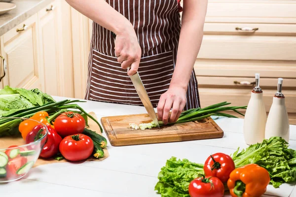 Mujer joven cocinando en la cocina en casa. Una mujer corta una cebolla verde y verduras con un cuchillo . — Foto de Stock