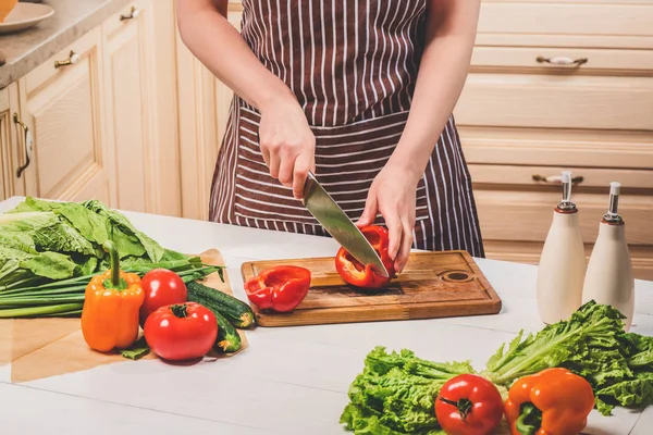 Mujer joven cocinando en la cocina en casa. Una mujer corta un pimiento y verduras con un cuchillo . — Foto de Stock
