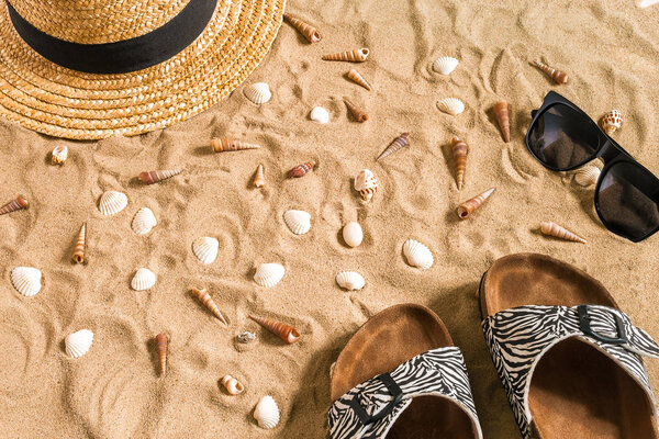 Summer beachwear, flip flops, hat, sunglasses and seashells on sand beach.