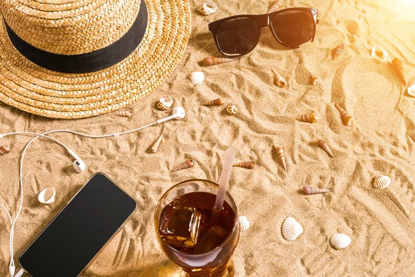 Summer beachwear, sunglasses, hat, cold drink in a glass and seashells on sand beach. — Stock Photo, Image