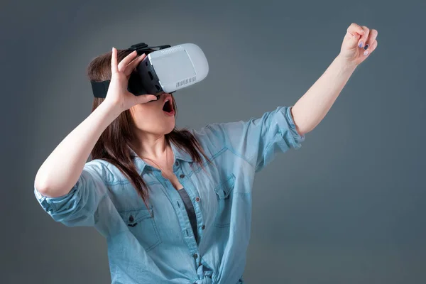 Giovane donna emotiva che utilizza un visore VR e sperimenta la realtà virtuale su sfondo grigio — Foto Stock