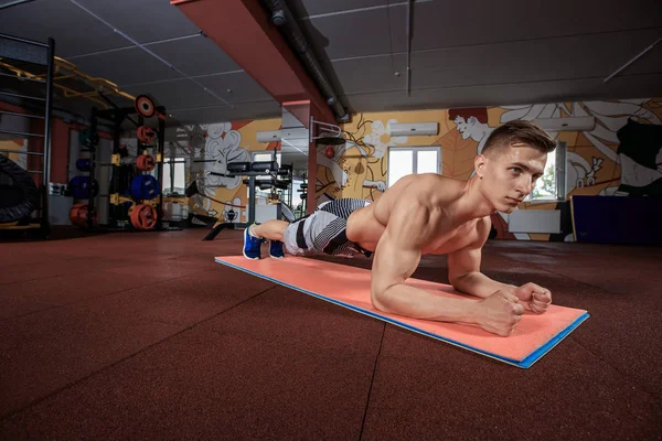 Kendine güvenen kaslı genç adam yerdeki spor salonunda egzersiz yaparken spor giyim ve iş tahta pozisyonu giyiyor
