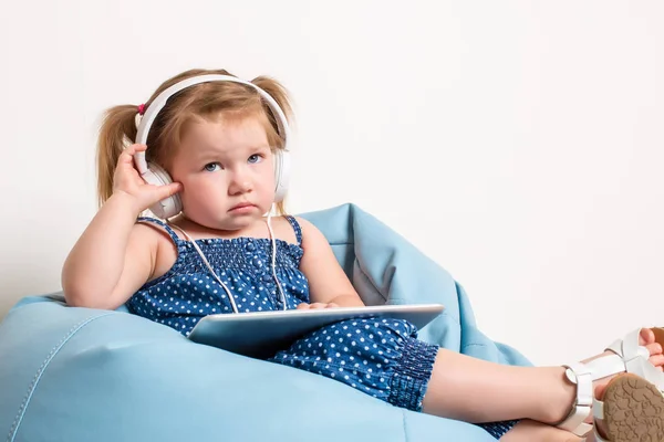 Милая маленькая девочка в наушниках слушает музыку с помощью планшета и улыбается, сидя на синей большой сумке — стоковое фото