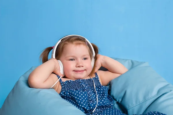 Menina bonito em fones de ouvido ouvindo música usando um tablet e sorrindo enquanto sentado em azul big bag — Fotografia de Stock