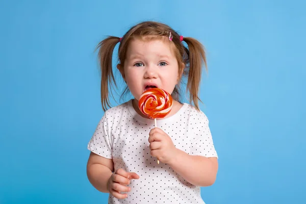 Szép kis női gyermek gazdaság hatalmas nyalóka spirál candy mosolygós boldog elszigetelt kék háttér. — Stock Fotó