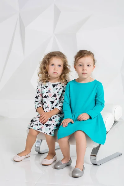Két kis barátnő azonos a ruhák, a különböző színek, ül egy széken, egy stúdióban, fehér falak — Stock Fotó