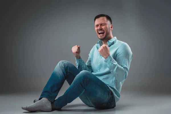 El hombre está sentado en el suelo, aislado sobre un fondo gris. Hombre mostrando diferentes emociones . —  Fotos de Stock