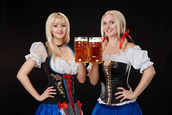 Две красивые блондинки держат в руках бокалы пива и стоят на черном фоне в студии . — стоковое фото