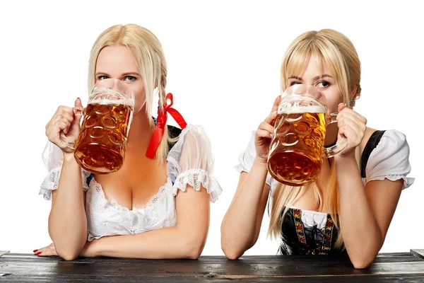 Zwei schöne Frauen trinken Bier aus großen Gläsern, während sie im Studio an einem Holztisch auf weißem Hintergrund sitzen — Stockfoto