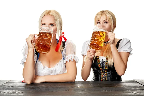 Дві красиві жінки п'ють пиво з великих окулярів, сидячи за дерев'яним столом на білому тлі в студії — стокове фото