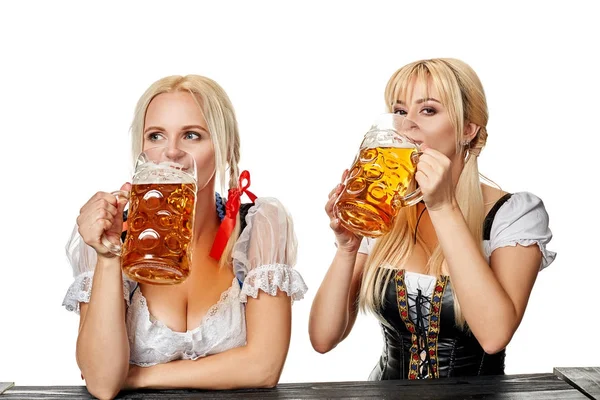 Zwei schöne Frauen trinken Bier aus großen Gläsern, während sie im Studio an einem Holztisch auf weißem Hintergrund sitzen — Stockfoto
