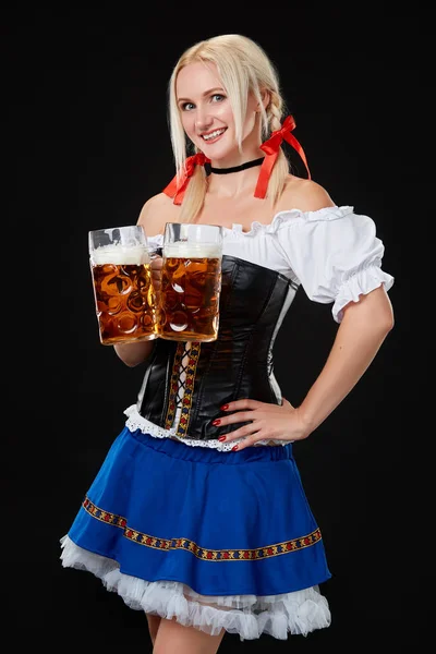 Junge sexy Frau trägt ein Dirndl mit zwei Bierkrügen auf schwarzem Hintergrund. — Stockfoto